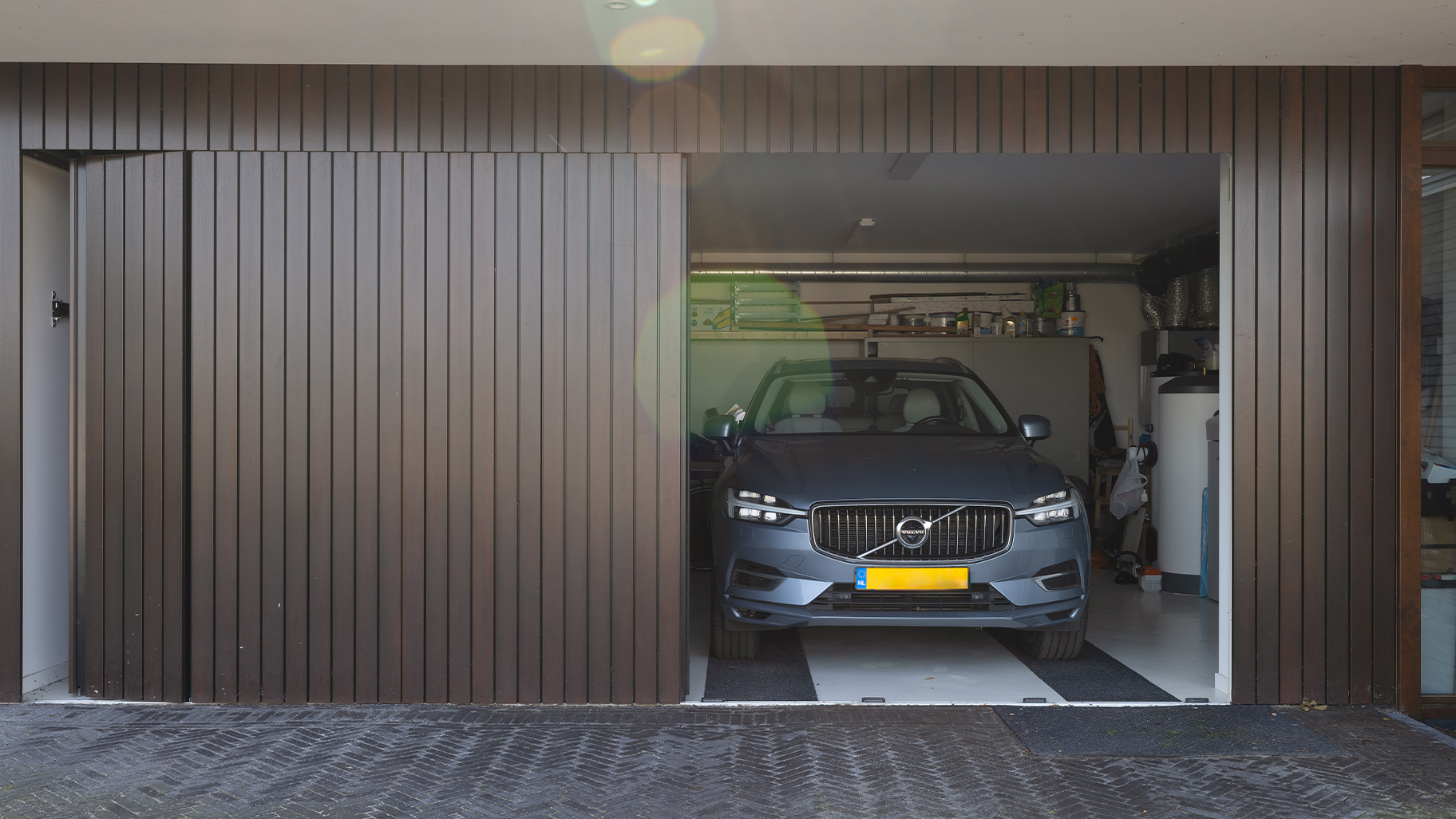 Onzichtbare zijwaartse garagedeur volledig gelijk is met de gevel geplaatst. Hierdoor lijkt de deur, wanneer gesloten, onzichtbaar en vormt deze een perfect doorlopende houten wand. De verticale houten latten geven de gevel een moderne, strakke uitstraling.