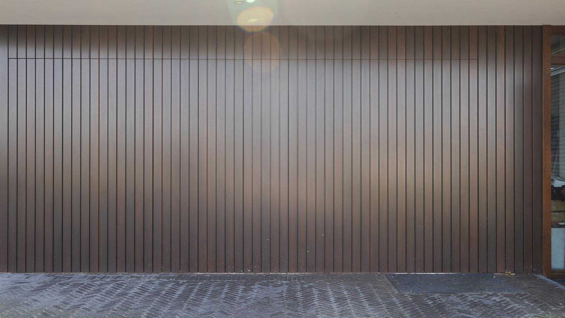 Onzichtbare zijwaartse garagedeur volledig gelijk is met de gevel geplaatst. Hierdoor lijkt de deur, wanneer gesloten, onzichtbaar en vormt deze een perfect doorlopende houten wand. De verticale houten latten geven de gevel een moderne, strakke uitstraling.