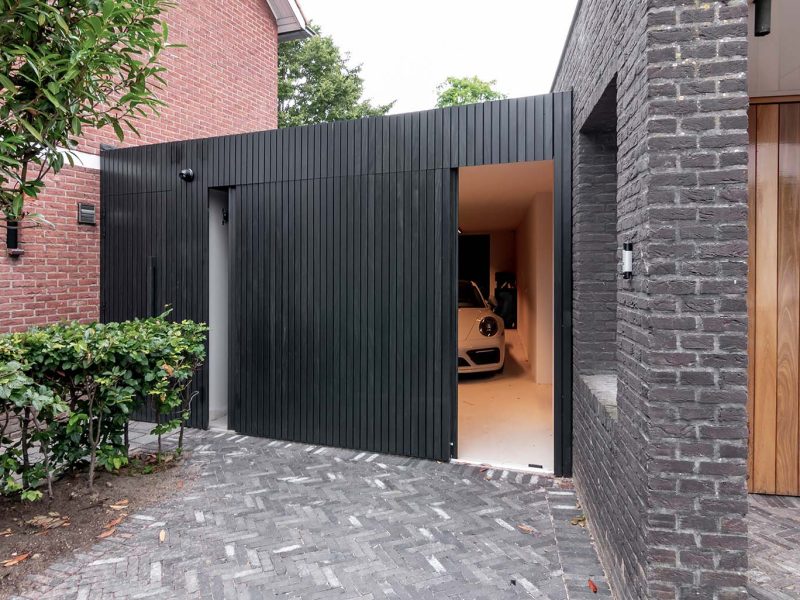 Moderne garage met een zwarte zijwaartse garagedeur van Afrormosia hout met een Porsche