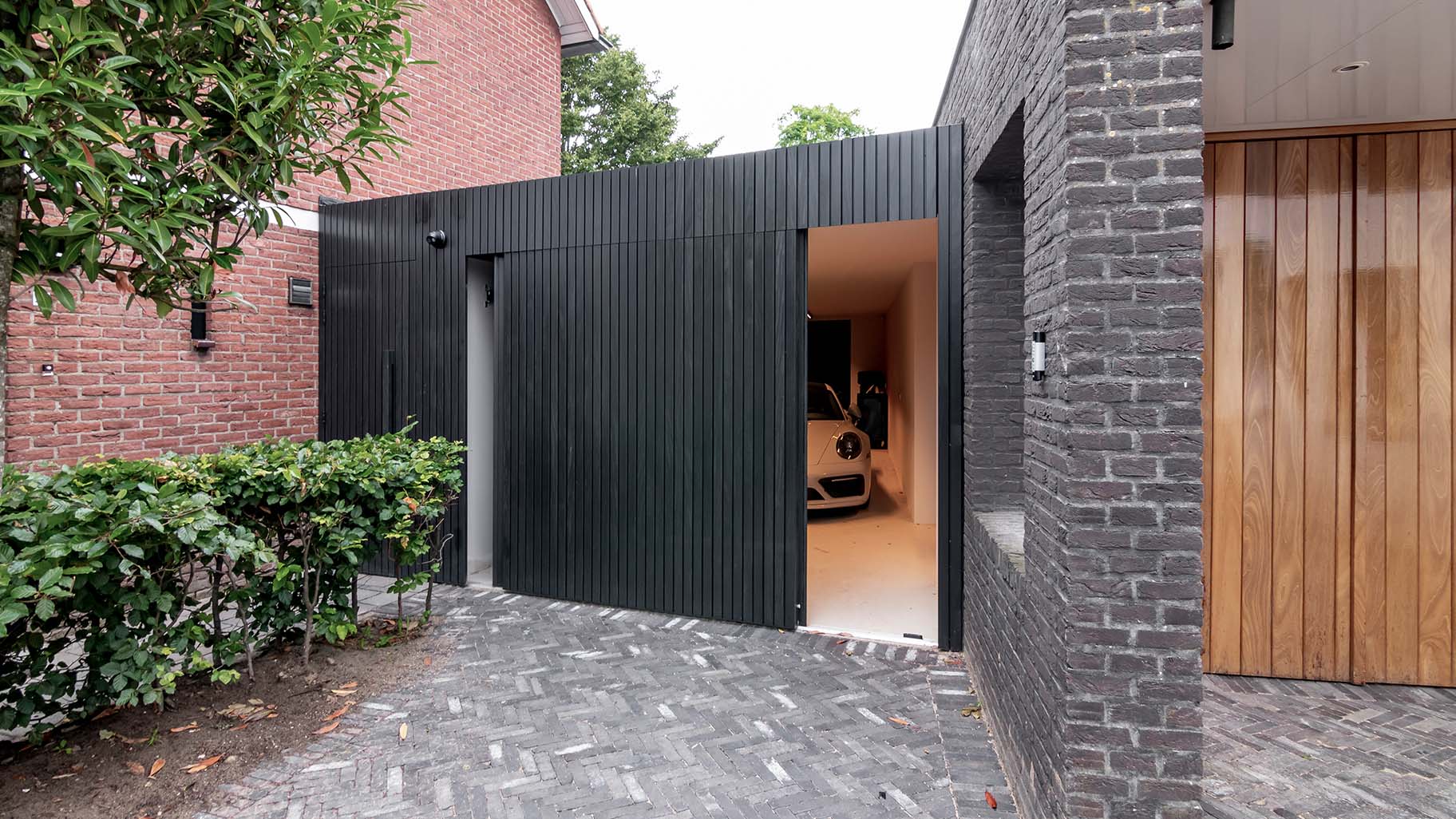 Moderne garage met een zwarte zijwaartse garagedeur van Afrormosia hout met een Porsche